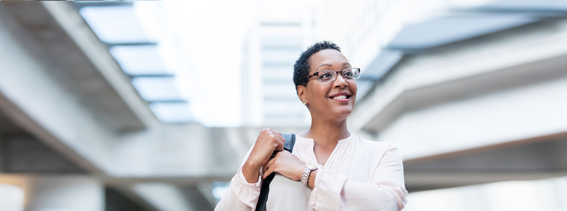 A mature African-American woman in her 40s walking on a city street, wearing eyeglasses and carrying a shoulder bag. She is in the foreground and the street scene behind her is out of focus.